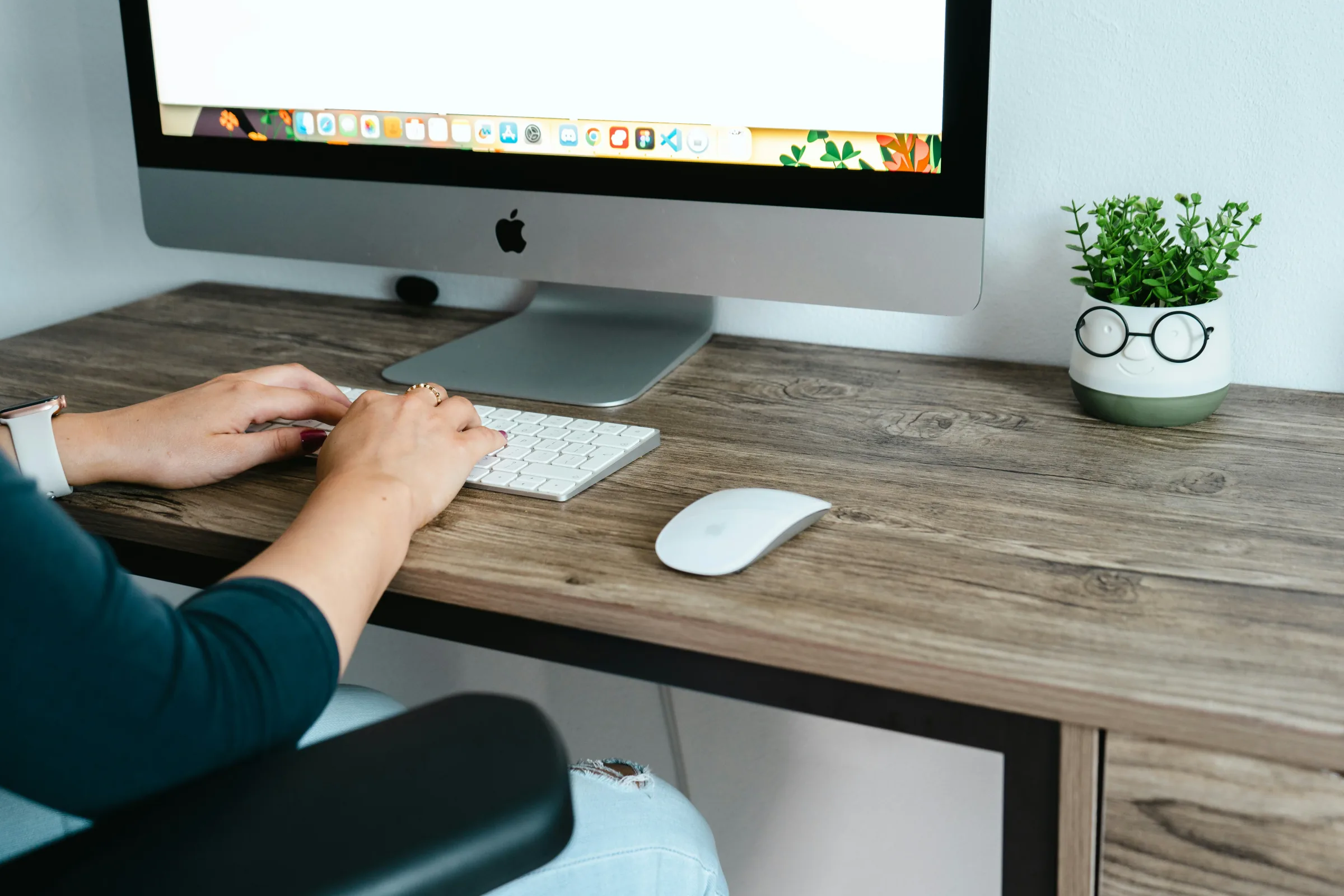 Mains en train de saisir au clavier d'un ordinateur de bureau, sur un plan de travail en bois avec une plante, poste de travail dans un environnement tertiaire.